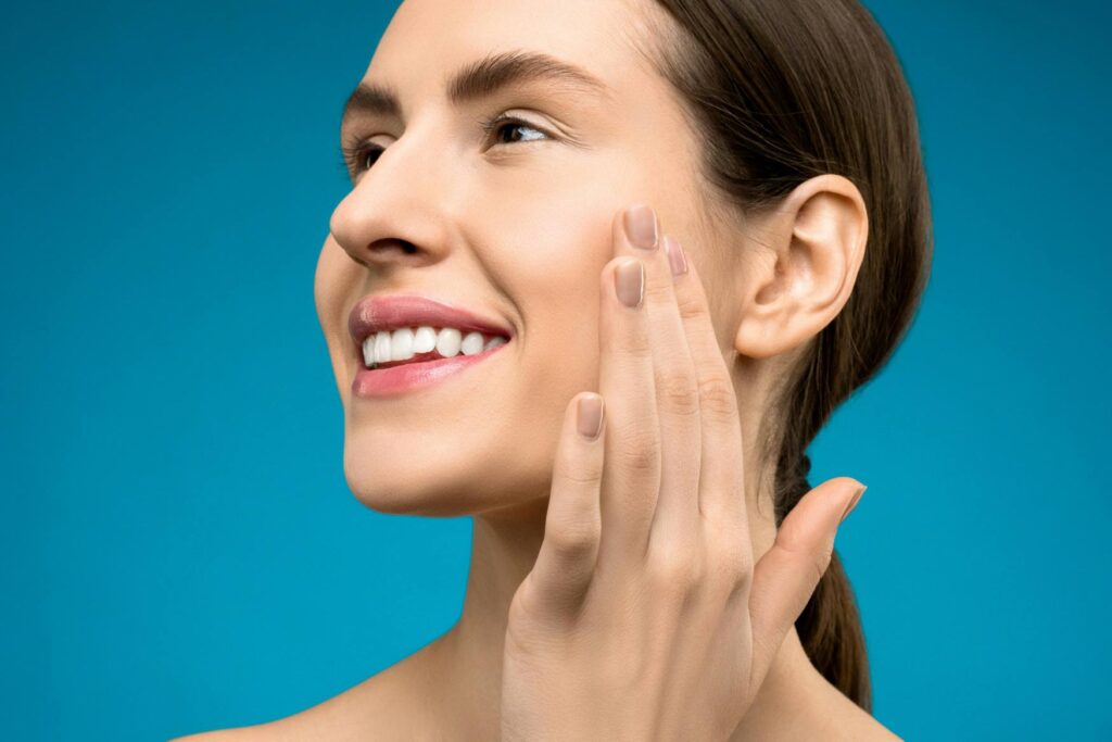 Woman smiling and touching her cheek with her hand against a plain blue background.