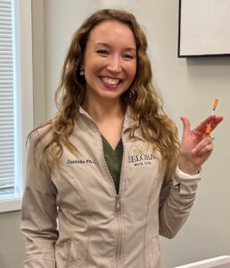 Smiling woman in a beige medical jacket holds a small orange syringe in her right hand inside a clinic room.