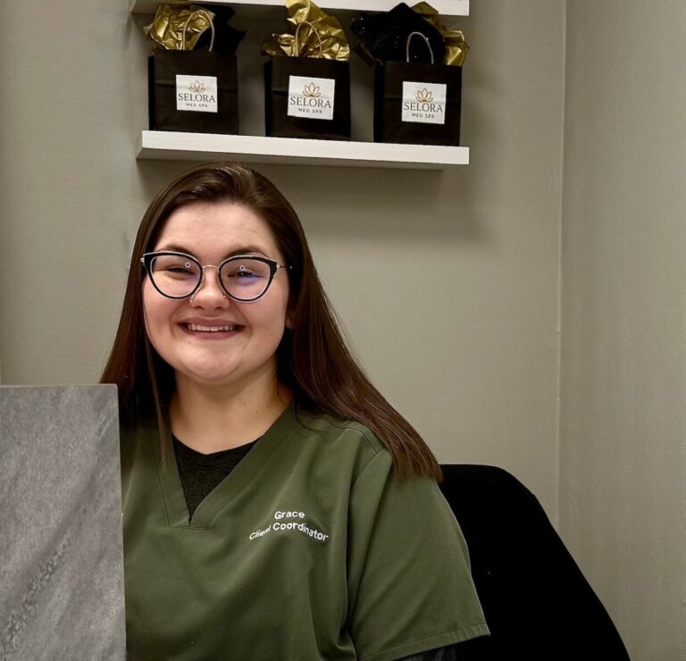 Woman wearing glasses and green scrubs sits at a desk, smiling, with gift bags on a shelf in the background.