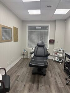 A medical exam room with a black reclining chair, side tables with supplies, and a window with closed blinds.
