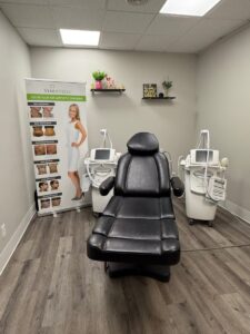 A treatment room with a black recliner chair, two medical devices, and a promotional banner for Venus Versa.