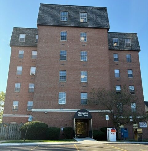 A five-story brick building labeled "Caldwell Building" with an arched entrance and a tree in front.