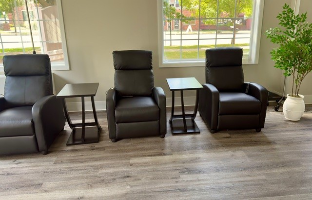 Three black recliner chairs with side tables are arranged in a row by large windows, with a potted plant on the right.