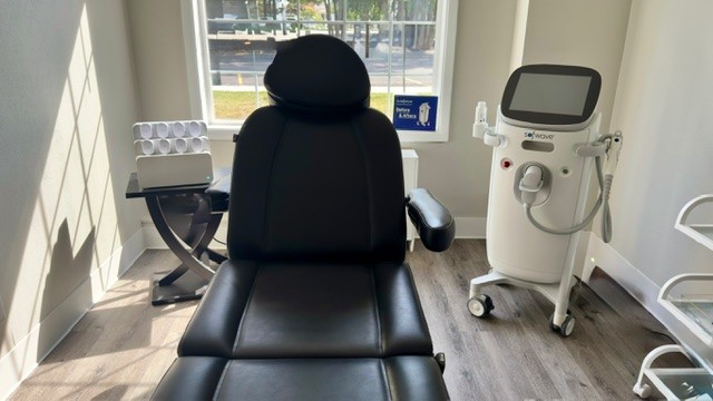 A black medical chair in a well-lit clinic room with equipment and a window in the background.