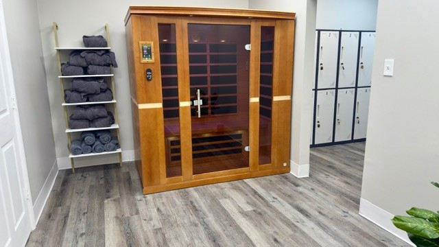A wooden sauna sits in a clean room with towel shelves on the left and lockers in the background.