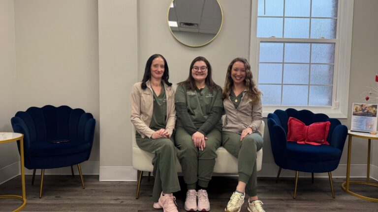 Three women in green scrubs sit on a white couch in a modern waiting room with navy chairs and a round mirror.