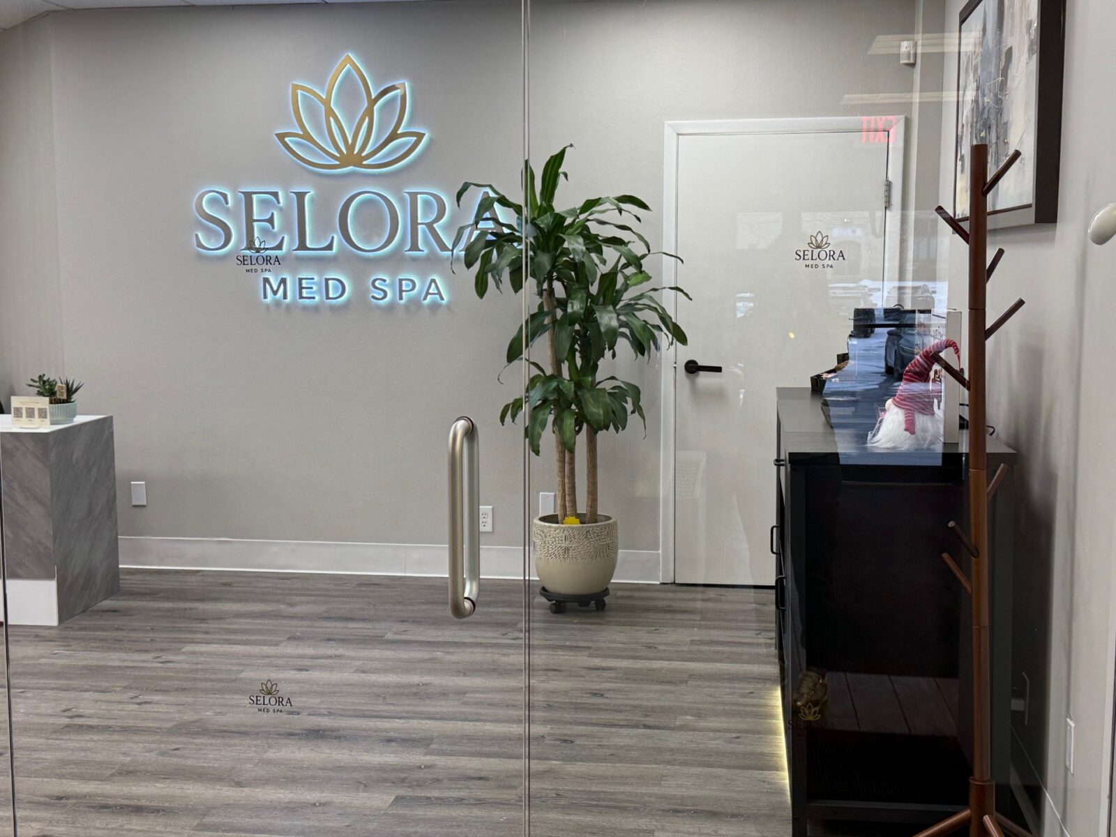 Reception area of Selora Med Spa with a potted plant, counter, coat rack, and glass door displaying the spa's logo.