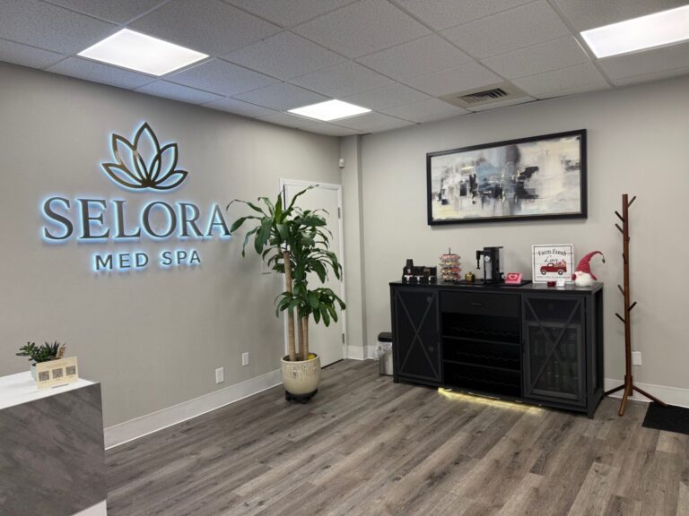 Reception area of Selora Med Spa with a plant, drink station, artwork, coat rack, and illuminated wall sign.